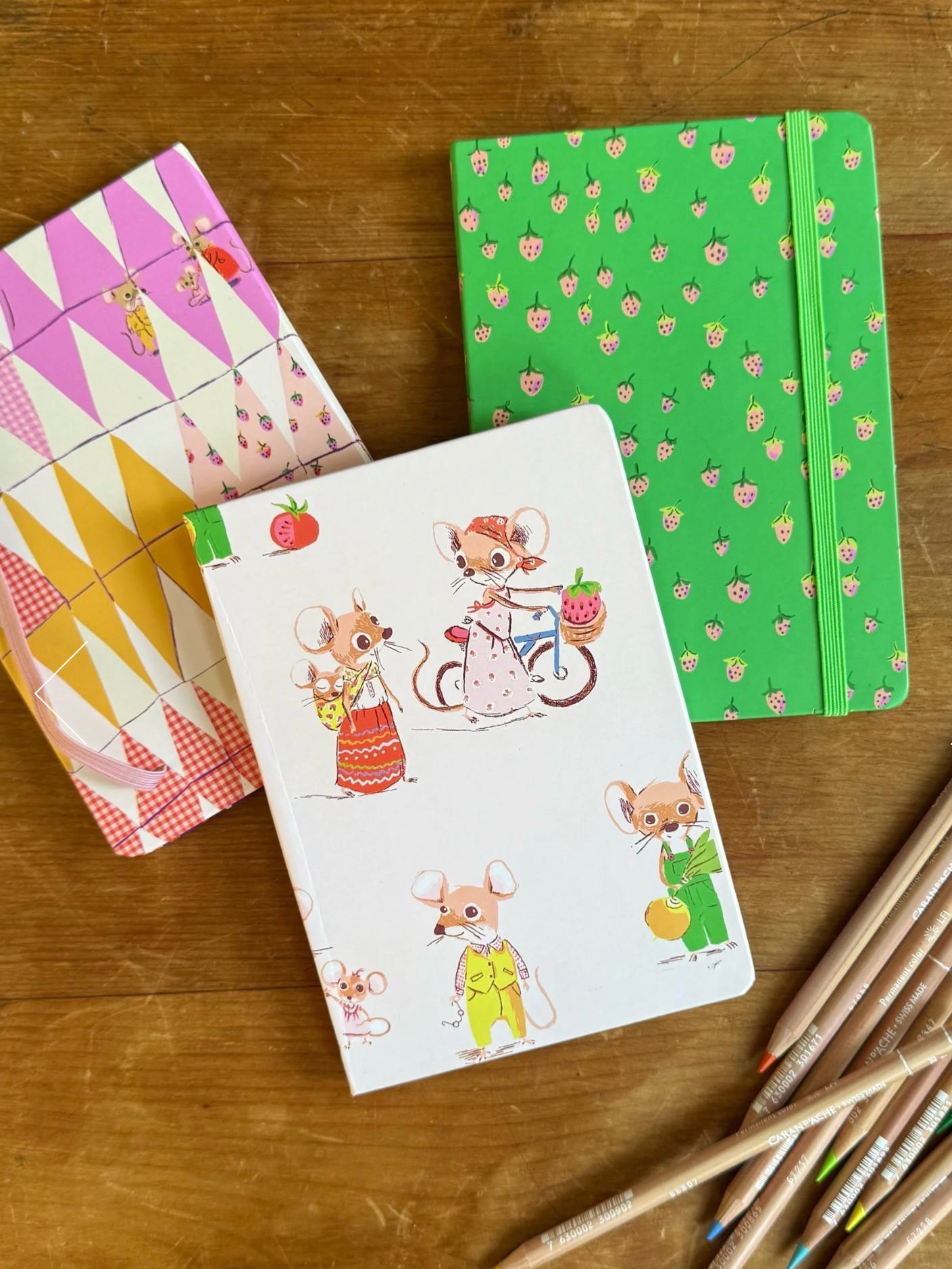 Three colorful notebooks with cartoon designs on a wooden surface, accompanied by colored pencils.